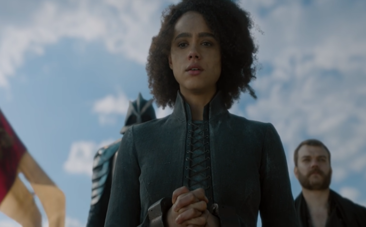 A screenshot of Game of Thrones, Season 8 (2019) that depicts Missandei before her execution. She is looking down towards the camera, which is angled up to show her and the sky. Her hands are bound in chains.