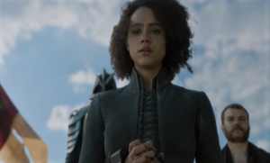 A screenshot of Game of Thrones, Season 8 (2019). It depicts Missandei before her execution. She is looking down towards the camera, which is angled up to show her and the sky. Her hands are bound in chains.